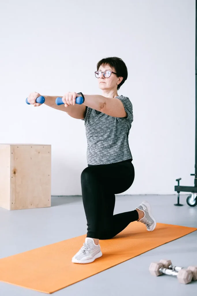 Người phụ nữ đeo kính quỳ một gối trên thảm cam, hai tay giữ tạ đơn nhỏ