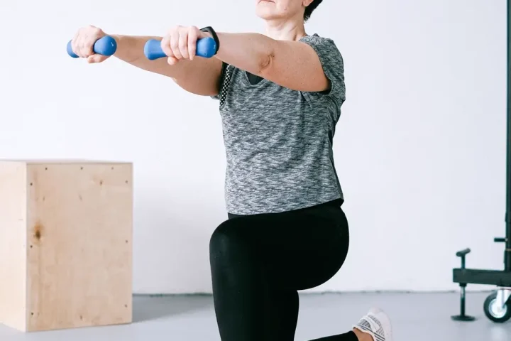Người phụ nữ đeo kính quỳ một gối trên thảm cam, hai tay giữ tạ đơn nhỏ