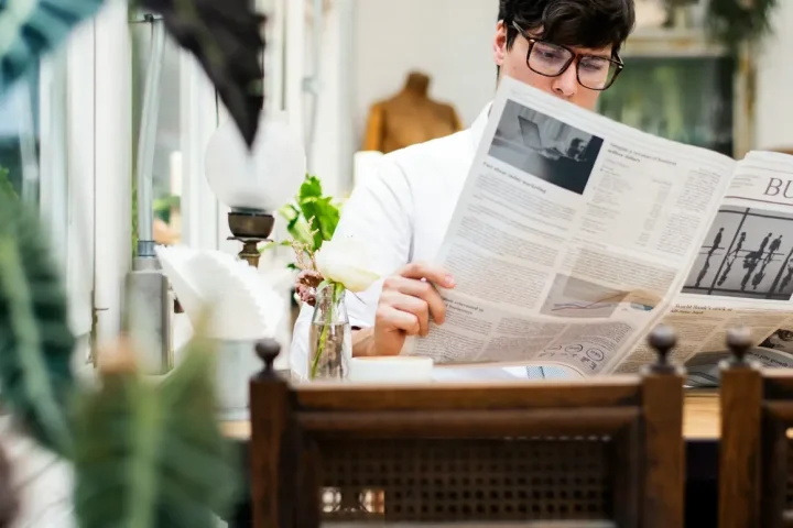 Người đàn ông đeo kính đọc báo giấy bên bàn gỗ cạnh cửa sổ