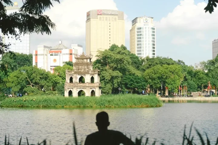 Tháp Rùa giữa hồ Gươm, hàng cây xanh và nhà cao tầng phía sau