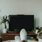 Phòng khách có ti vi, cây xanh và đôi chân gác lên bàn thư giãn